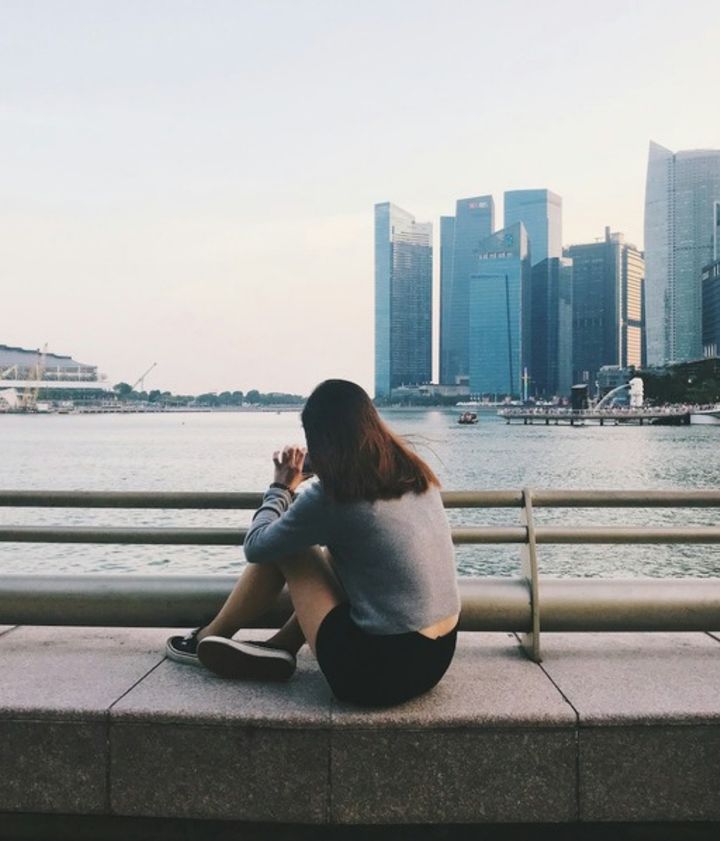 A young woman sitting alone by the water, facing the Singapore skyline, seen from behind